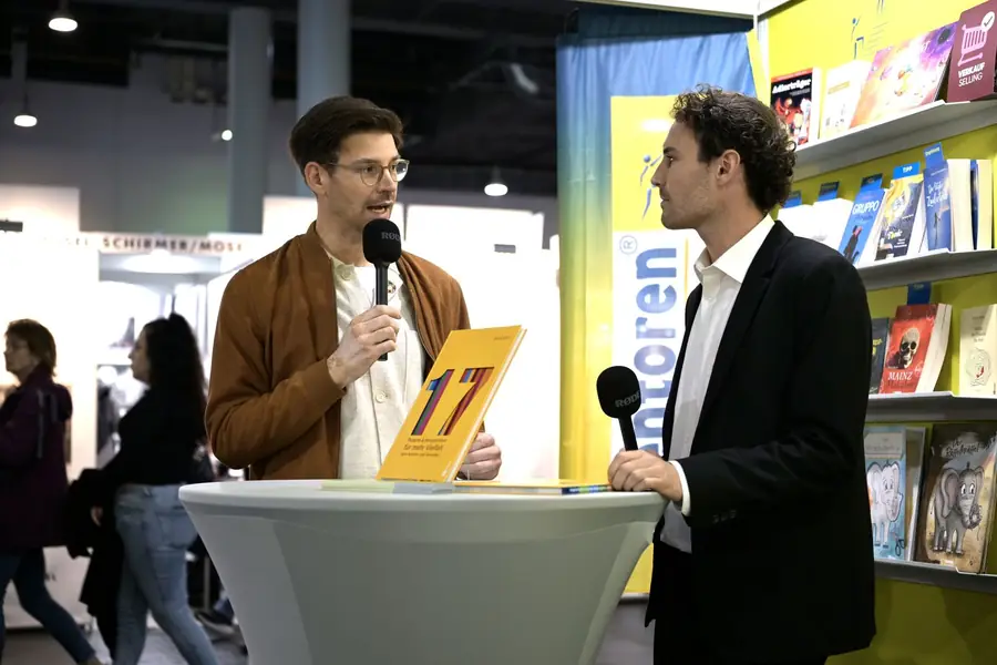 Bühnengespräch am Messestand: Jean Bauer mit Mikrofon und dem Buch „17“